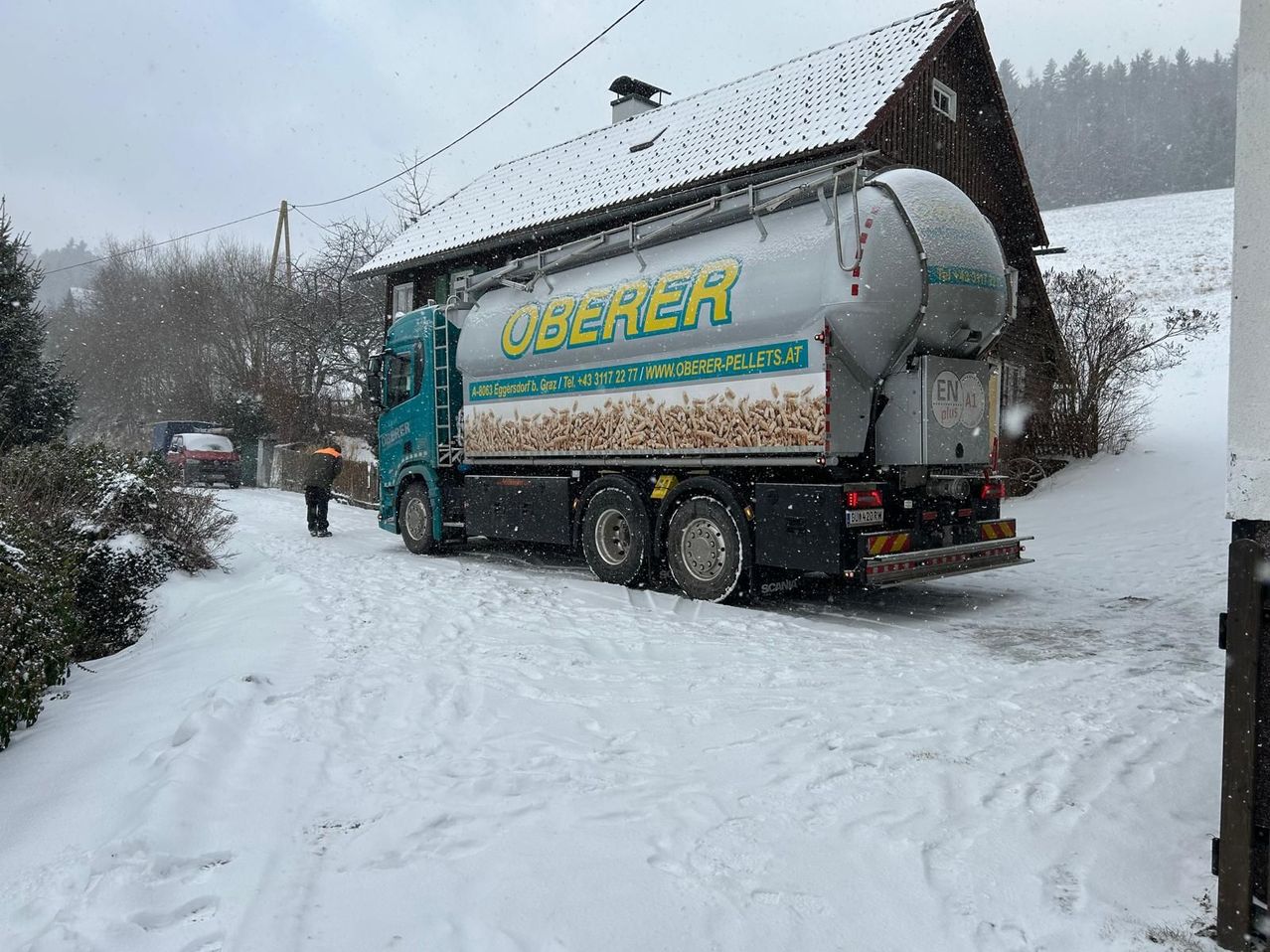 Ein LKW mit 'Oberer' drauf steht im Schnee neben einem Gebäude. Eine Person steht im Schnee in der Nähe.