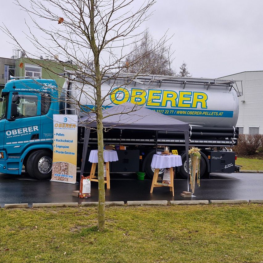 Ein blauer Lkw mit dem Namen Oberer ist auf der Straße geparkt. Vorne steht ein Vordach mit zwei Tischen. Links befindet sich ein Banner mit demselben Namen.