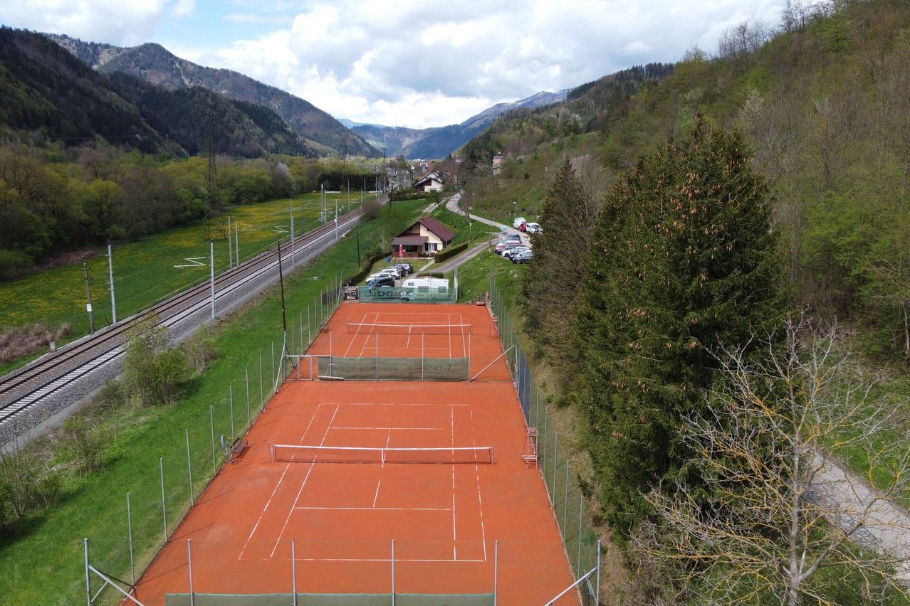 Luftaufnahme von Tennisplätzen in einem malerischen Tal mit Bergen, Häusern und einer Bahnstrecke. Die Plätze sind leer mit Netzen und Stangen.