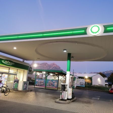 Eine BP-Tankstelle bei Dämmerung mit grünen und weißen Schildern, Zapfsäulen und einem Geschäft. Der Himmel ist blau mit Bergen im Hintergrund.