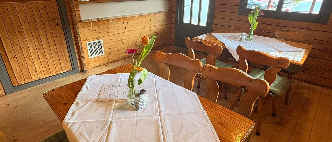 Ein holzverkleideter Essraum mit einem Tisch, der mit einem weißen Tuch bedeckt ist. Tulpen in einer Vase stehen auf dem Tisch. Der Raum hat Holzwände und -böden. Es gibt weitere Tische und Stühle. Links befindet sich eine Tür.