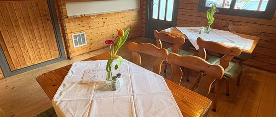 A wooden dining room with a table covered in white cloth. Tulips in a vase are on the table. The room has wooden walls and floors. There are other tables and chairs. There is a door on the left.