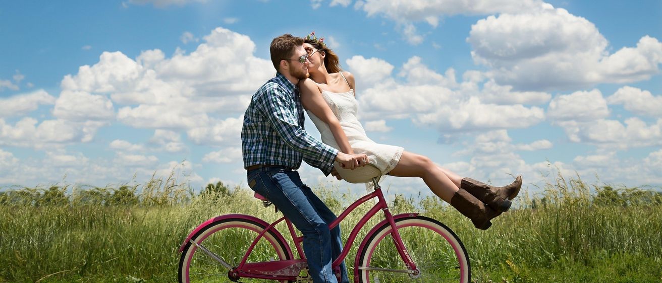 Ein Mann und eine Frau küssen sich auf einem rosa Fahrrad in einem Grasfeld unter einem blauen Himmel mit weißen Wolken.