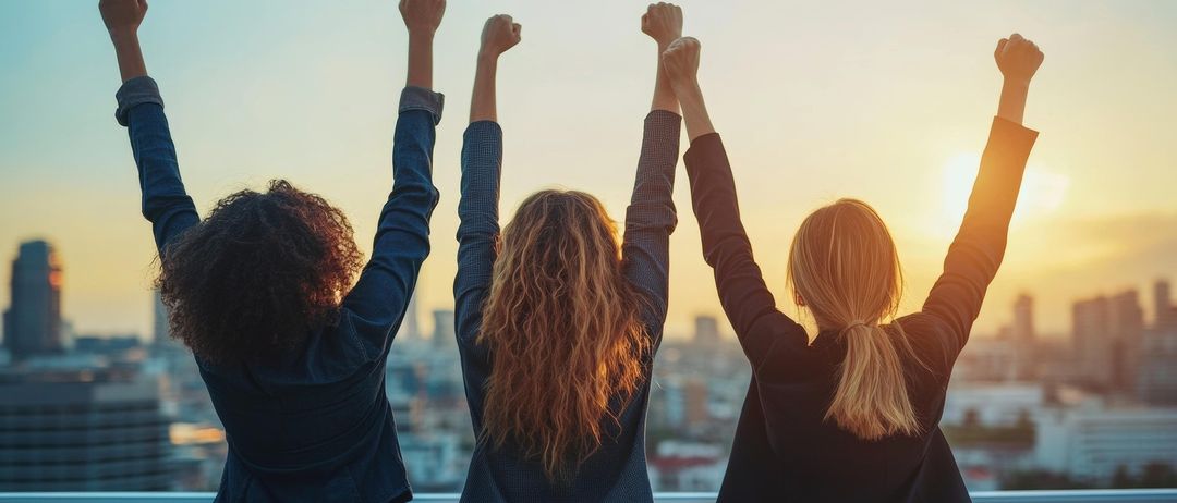 Drei Frauen stehen in einer Reihe mit erhobenen Armen und blicken auf eine Stadtlandschaft bei Sonnenuntergang.
