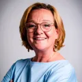 Eine Frau mit kurzen blonden Haaren und Brille posiert für ein Foto. Sie lächelt und trägt ein blaues Hemd und eine Halskette.
