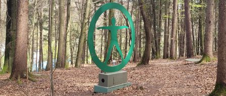 Eine grüne Skulptur einer menschlichen Figur steht prominent in einem dichten Wald, umgeben von kahlen Bäumen und einer Decke aus gefallenen Blättern.
