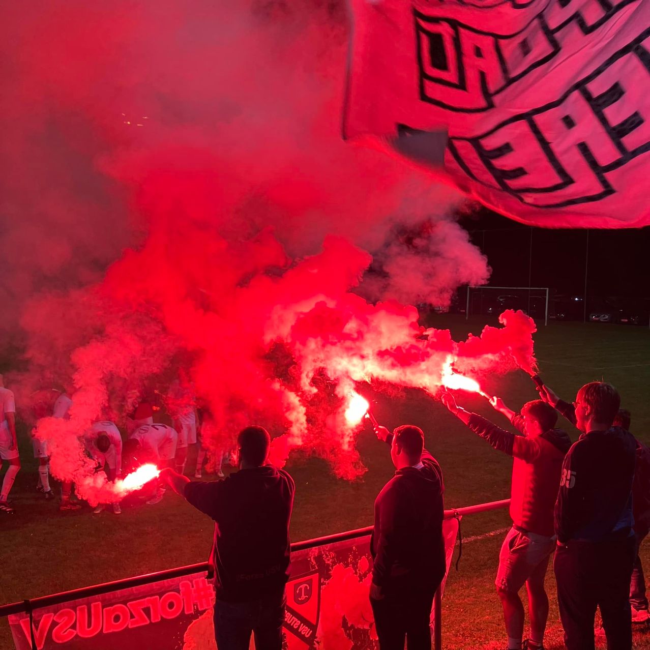 Fans bei einem Fußballspiel halten rote Fackeln und erzeugen eine dramatische Szene mit rotem Rauch, beleuchtet vom Glühen der Fackeln. Die Spieler sind im Hintergrund zu sehen.