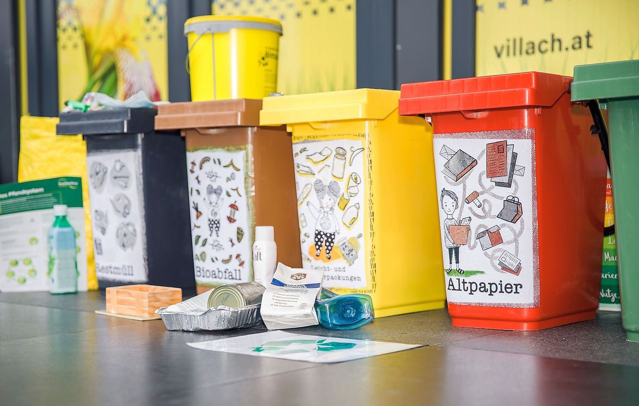 Ein Tisch mit verschiedenen farbigen Recycling-Behältern, darunter gelb für Bioabfall, braun für Altpapier und rot für andere Abfälle.