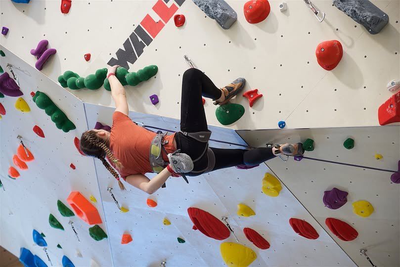 Eine Frau klettert an einer Kletterwand mit bunten Griffen. Sie trägt einen Helm, einen Gurt und Turnschuhe.