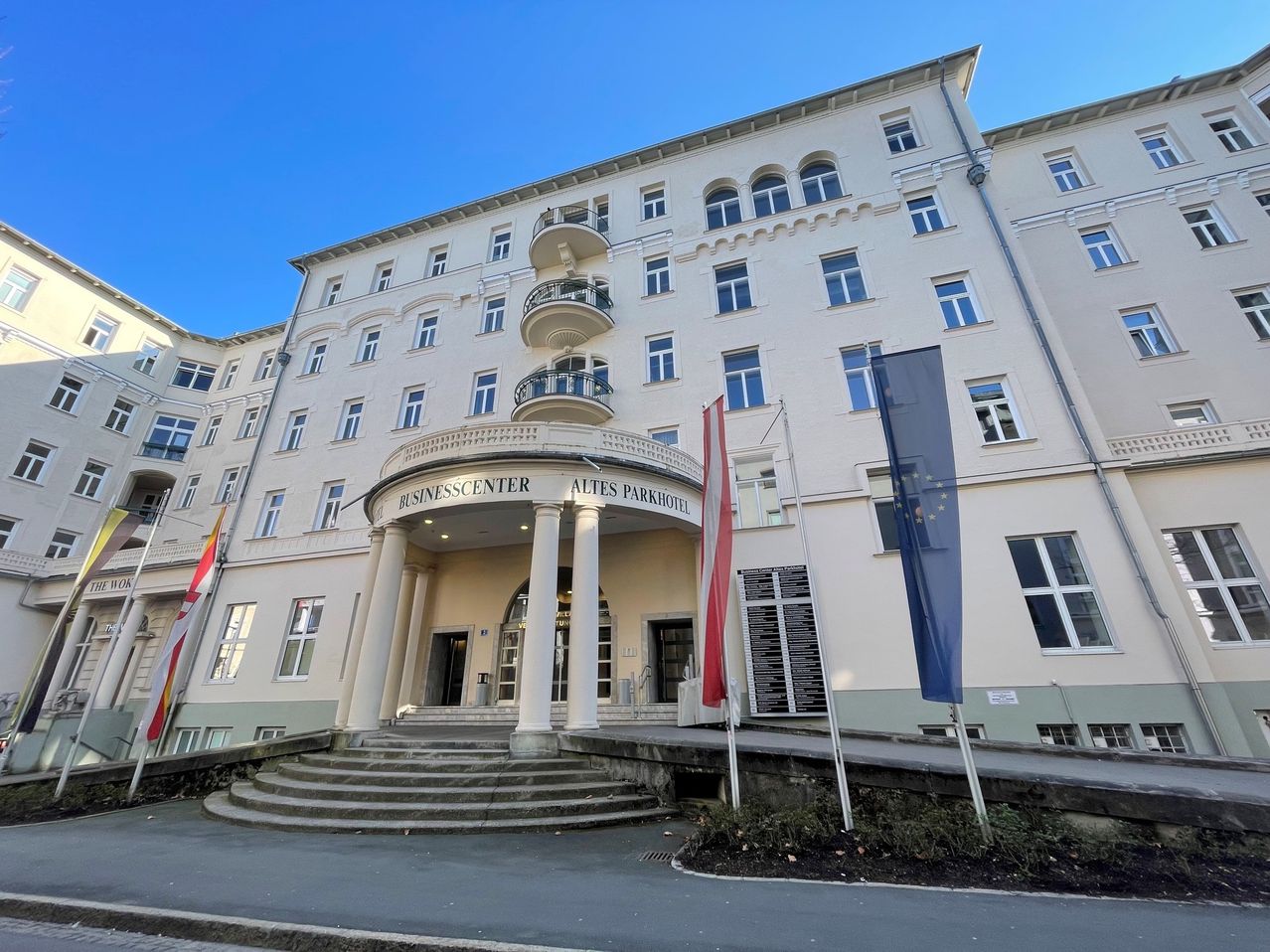 Das Altes Parkhotel Business Center verfügt über einen großen Eingang mit Säulen und einer Treppe. Zwei Fahnen, eine rot-weiß und die andere blau, sind an den Seiten angebracht.
