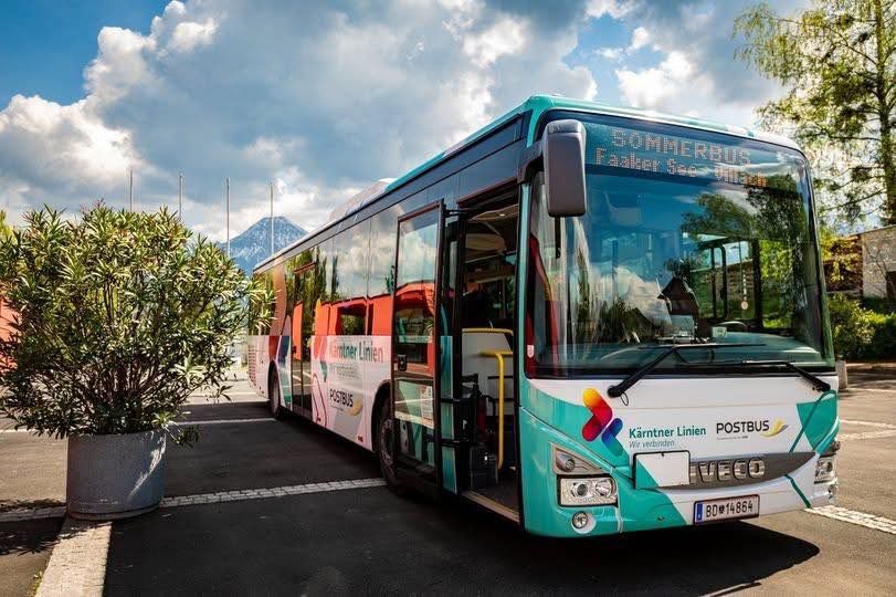 Ein grüner und weißer Bus mit dem Wort 'Sommerbus' auf der Vorderseite ist auf einem Parkplatz geparkt, mit einem Berg im Hintergrund.