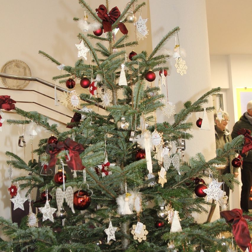 Ein Weihnachtsbaum, geschmückt mit roten Kugeln, weißen Schneeflocken und Schleifen, steht in einem Raum. Er befindet sich in der Nähe einer Treppe und einer weißen Wand. Zwei Personen stehen dahinter.