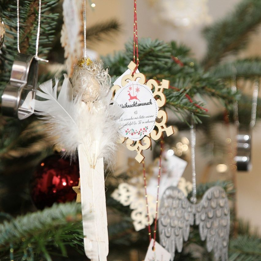 Ein Weihnachtsbaum mit verschiedenen Ornamenten, darunter ein roter Ball, weiße Federn, Engelsflügel und ein holzfarbener Schneeflocke. Die Schneeflocke trägt die Aufschrift 'Leichnachtswunsch' und wird von einer kleinen Engelsfigur begleitet.