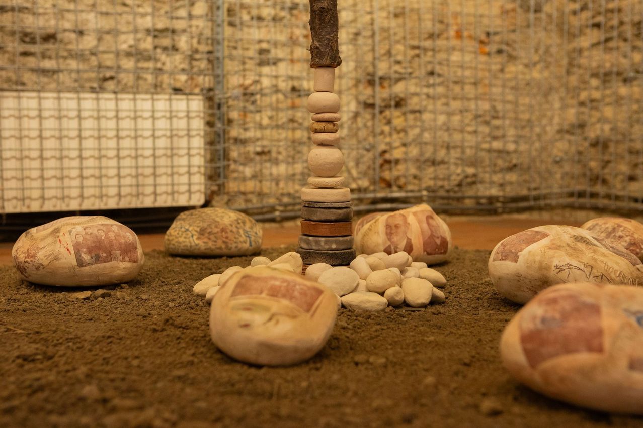 Eine Sammlung von Steinen, einschließlich eines Stapels von Kieselsteinen und Steinen mit Gesichtern, ist auf einer Erdoberfläche angeordnet. Ein Metallzaun und eine weiße Wand sind im Hintergrund.