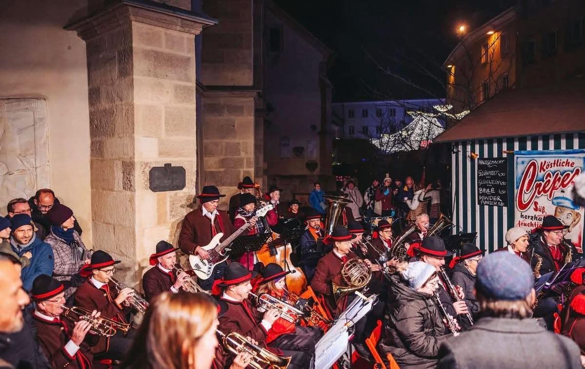Ein nächtliches Konzert einer Blechbläser-Band und eines Gitarristen vor einem Publikum. Einige Musiker sitzen, andere stehen. Sie tragen einheitliche Kleidung. Der Auftritt findet im Freien statt, mit Gebäuden und Bäumen im Hintergrund.