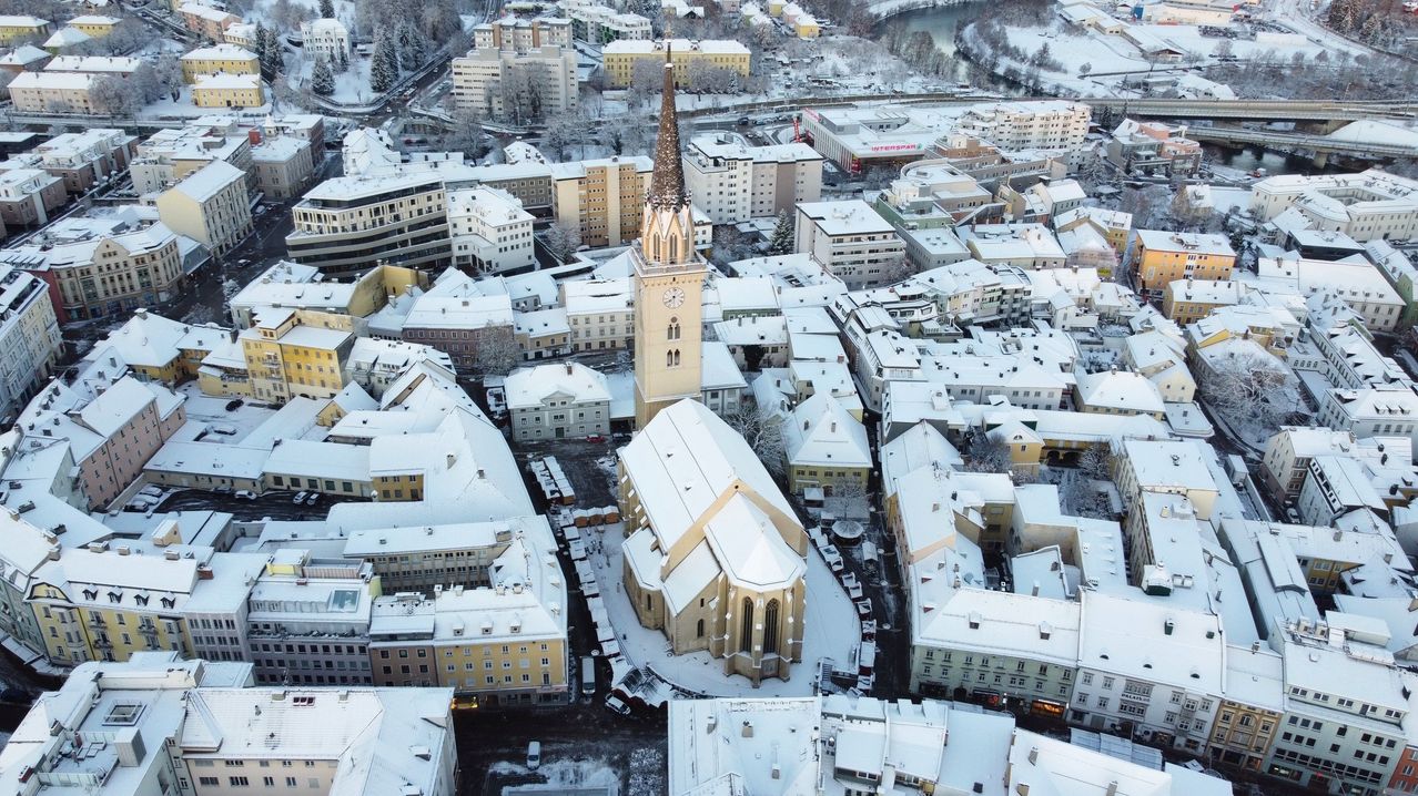 Luftaufnahme einer schneebedeckten Stadt mit einem hohen Kirchturm. Die Stadt zeigt verschiedene Gebäude, einige mit schneebedeckten Dächern. Ein Fluss fließt durch das Gebiet, und ein großes Gebäude mit dem Wort Interspar ist sichtbar.