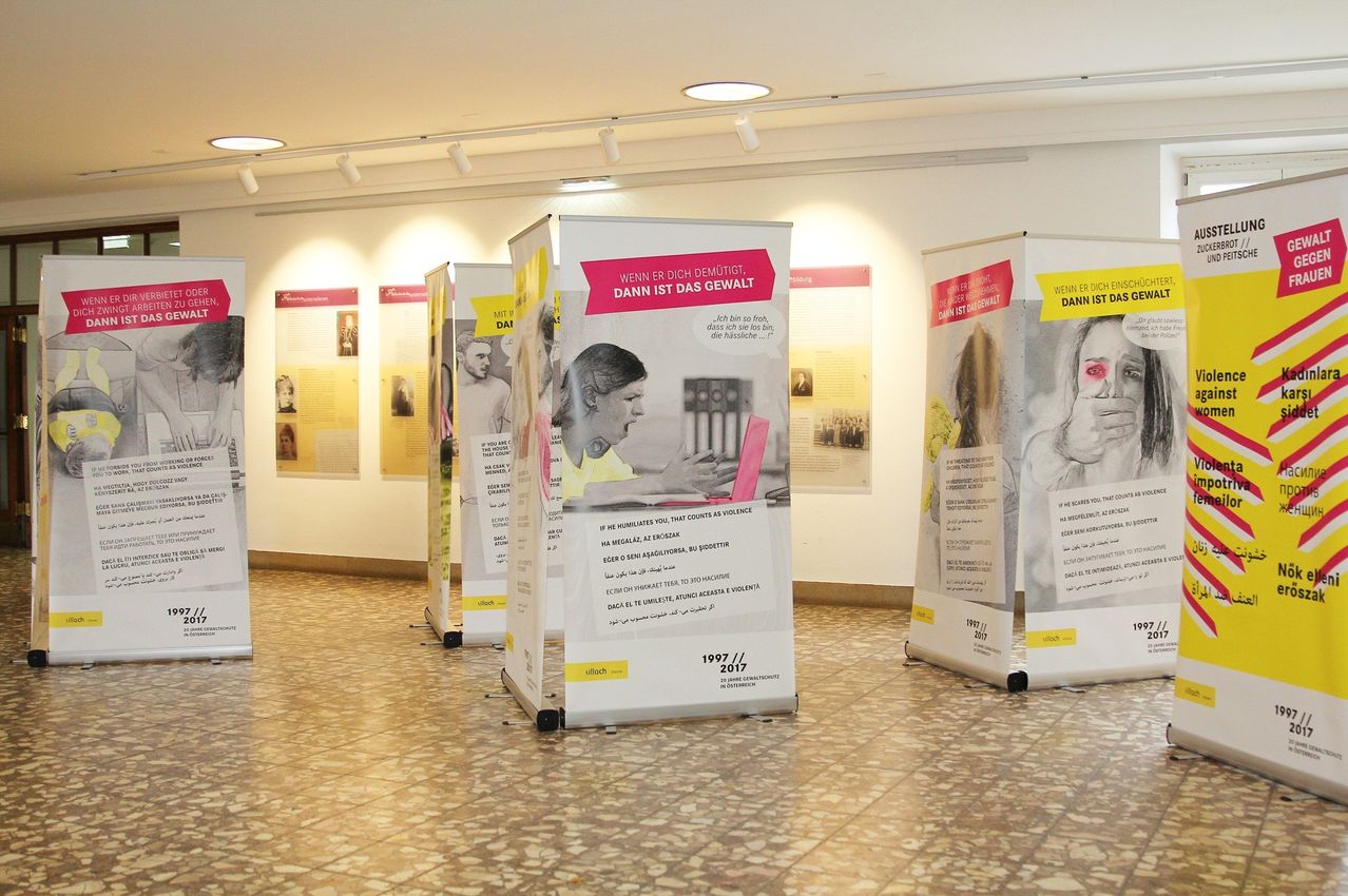 Ein Raum mit gefliesten Böden hat Poster und Banner mit Bildern und Text. Die Poster haben Bilder von Menschen, und die Banner haben ein Frauengesicht mit roten Augen. Der Raum hat weiße Wände und mehrere Lampen an der Decke.