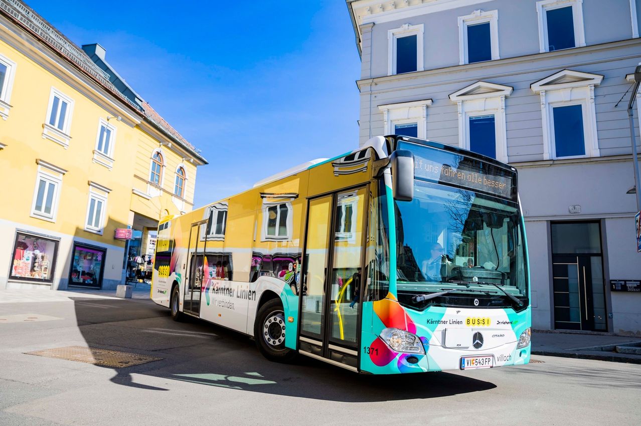 Ein großer mehrfarbiger Bus mit den Worten 'Kärtner Linien' fährt vor einem Gebäude mit vielen Fenstern auf einer Straße.