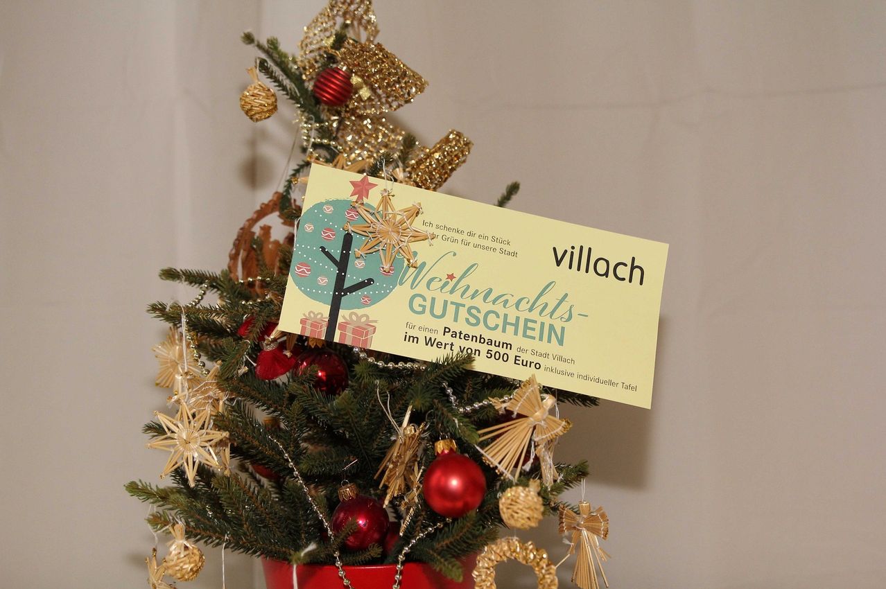 Ein geschmückter Weihnachtsbaum mit einem Schild, das einen Geschenkgutschein im Wert von 500 Euro aus der Stadt Villach anbietet. Der Gutschein ist gelb mit Stern- und Schleifendesign.