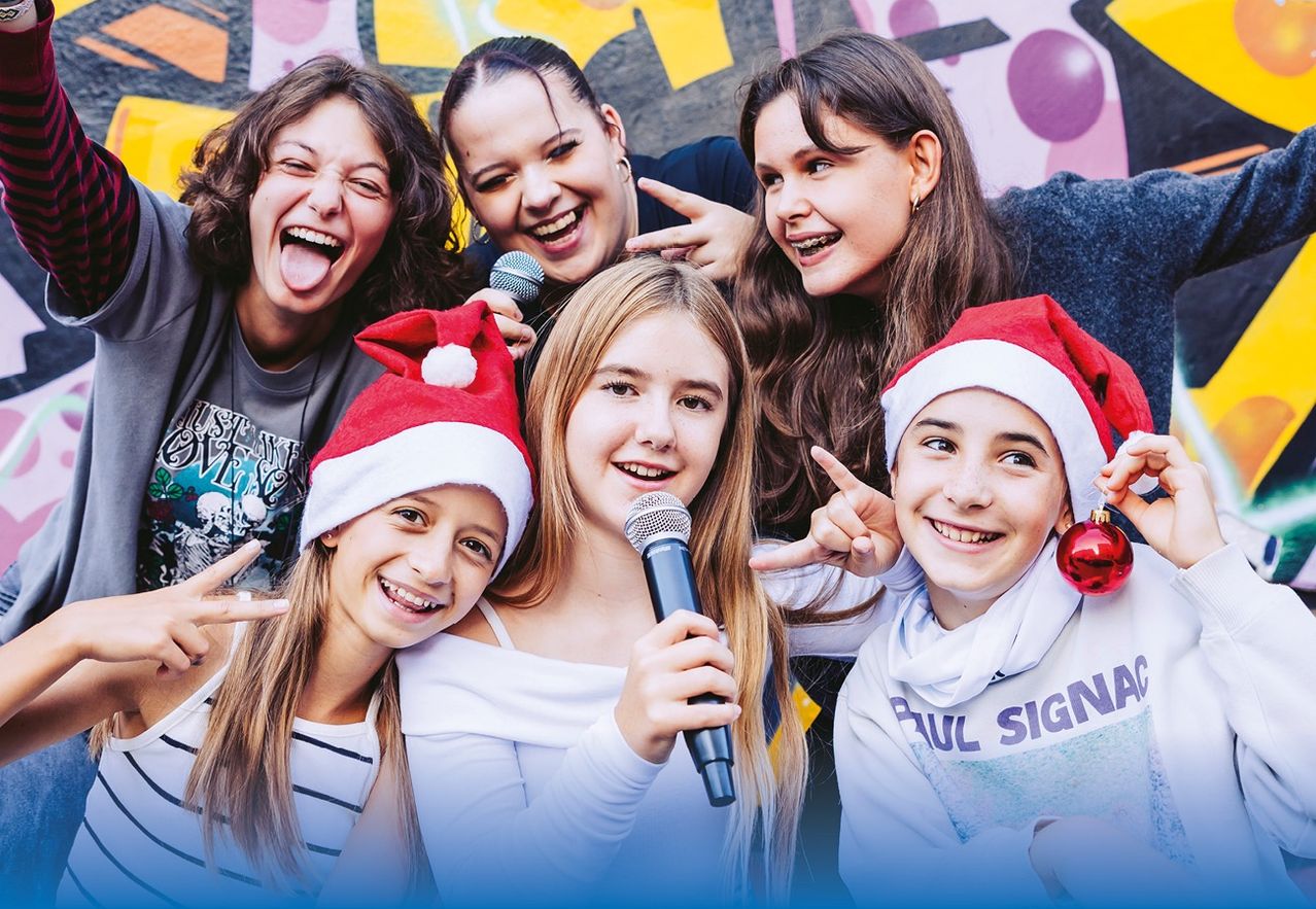 Eine Gruppe junger Mädchen mit Weihnachtsmützen und breitem Lächeln bei einer Fotoaufnahme. Eines der Mädchen hält ein Mikrofon und andere posieren mit Friedenszeichen und einer Zeigegeste. Sie scheinen auf einer Weihnachtsfeier zu sein.