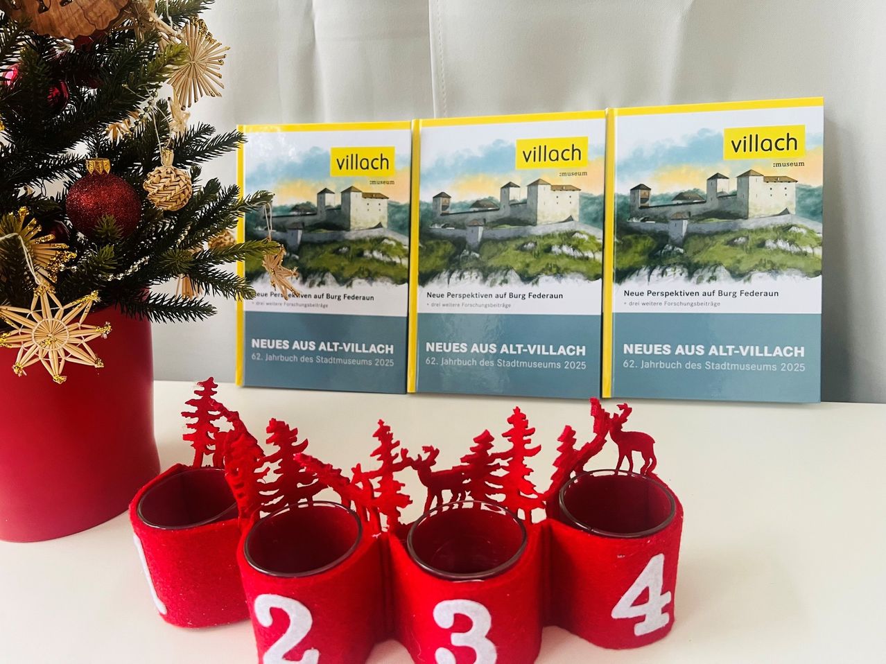 Drei Bücher mit dem Titel 'Neues aus Alt-Villach' und vier rote Kerzenhalter mit den Nummern 2, 3 und 4, neben einem Weihnachtsbaum und einer Vase auf einer weißen Oberfläche.
