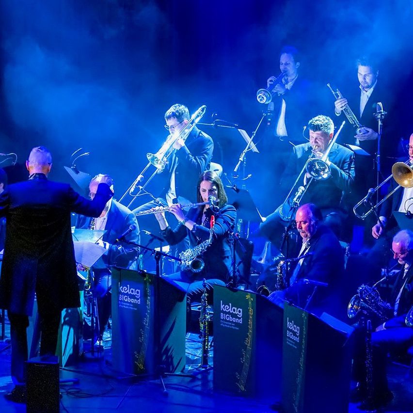 Eine Jazzband tritt auf der Bühne bei blauem Licht auf, mit Posaunen, Saxophonen und einem Dirigenten.