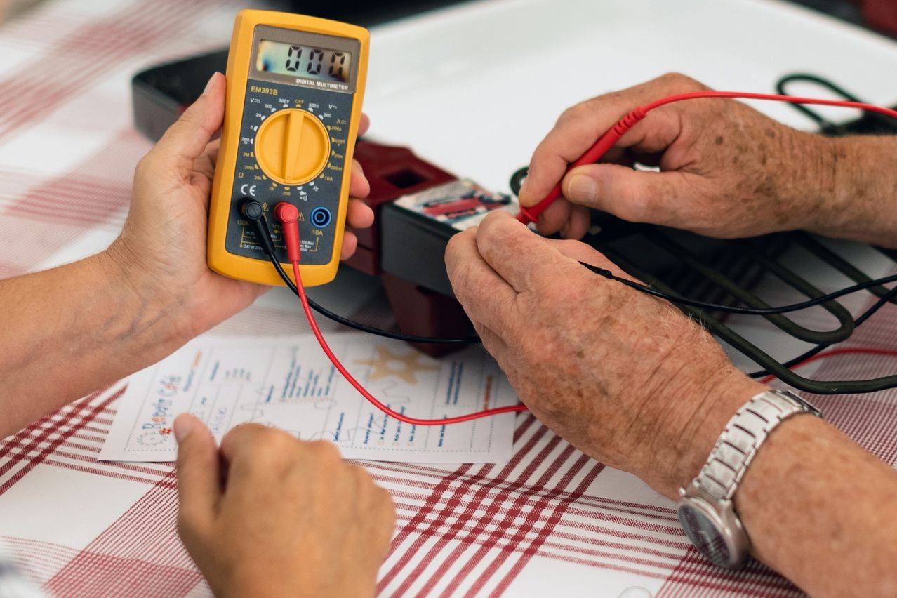 Eine Person hält ein digitales Multimeter und testet Kabel mit einer roten Sonde, während eine andere Person die schwarze Sonde hält.