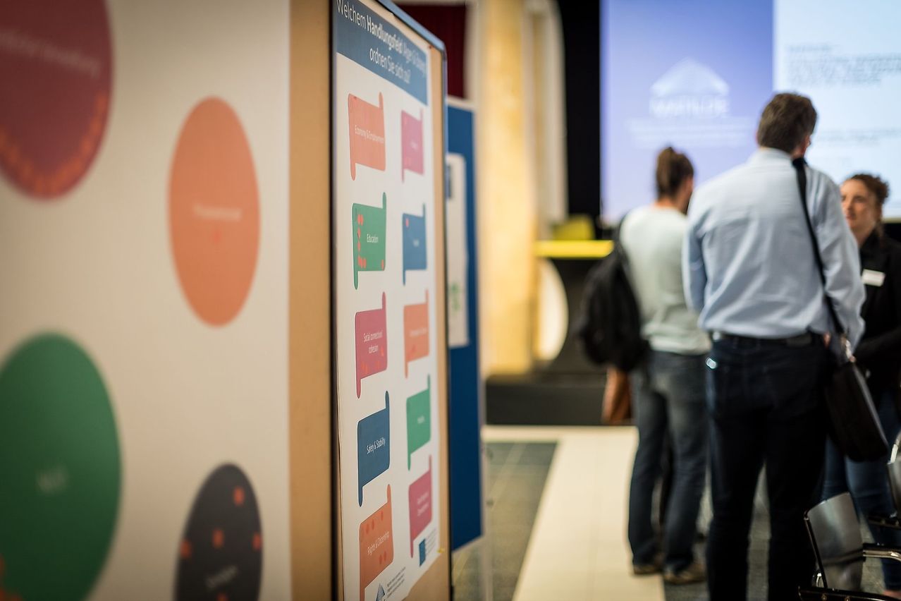 Zwei Personen stehen vor einem großen Posterboard mit verschiedenen farbigen Abschnitten und Text, ein unscharfer Projektionsbildschirm dahinter.