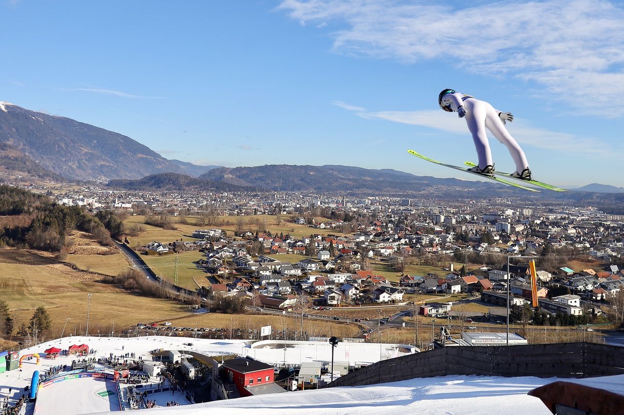 Ein Skifahrer schwebt in der Luft über einer verschneiten Landschaft, mit einer Stadt und Bergen im Hintergrund, unter einem teilweise bewölkten Himmel.