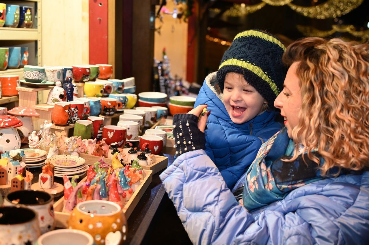 Ein junges Kind und eine erwachsene Frau sind bei einem Stand, lächeln und haben viele bunte Tassen und Figuren vor sich.