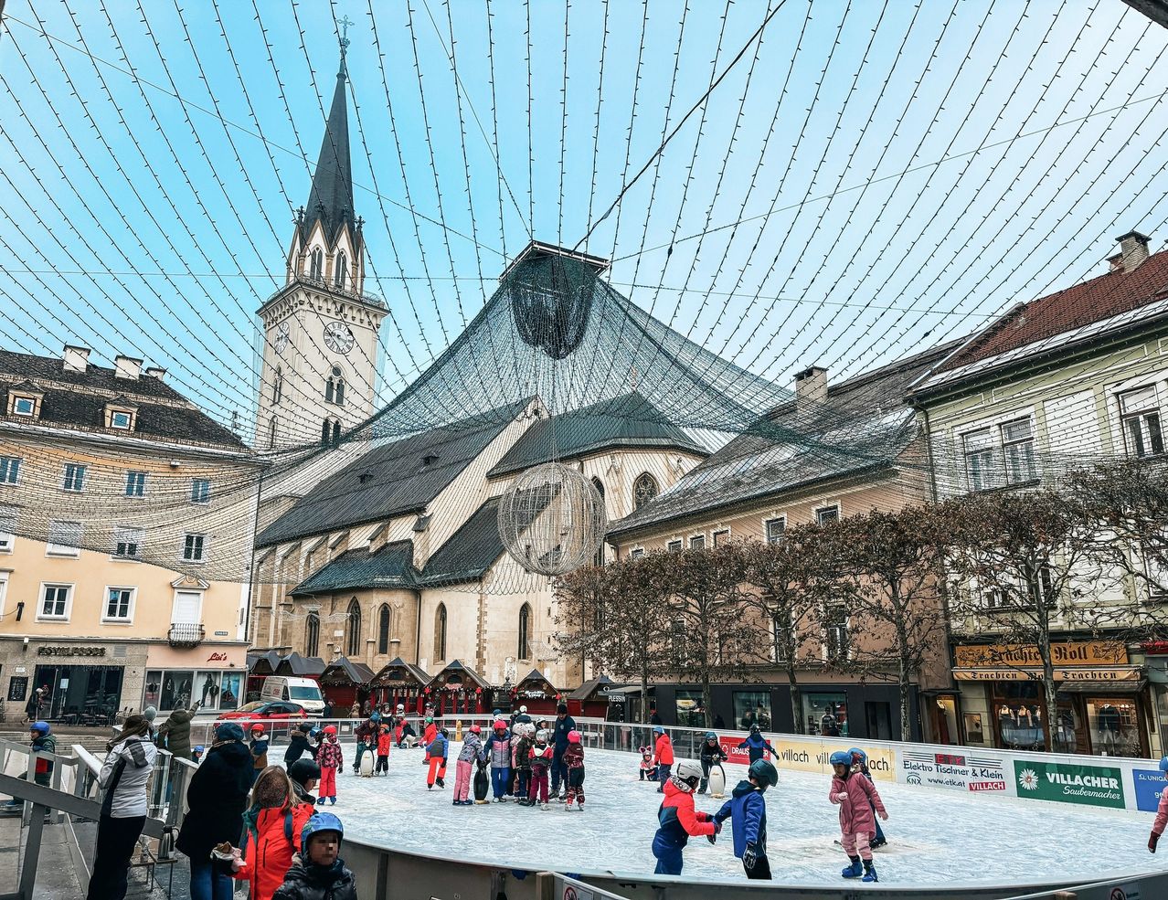 Eine Eisbahn auf dem Stadtplatz mit Schlittschuhläufern. Dahinter steht eine Kirche mit Turm und Spitzhelm. Der Himmel ist klar.