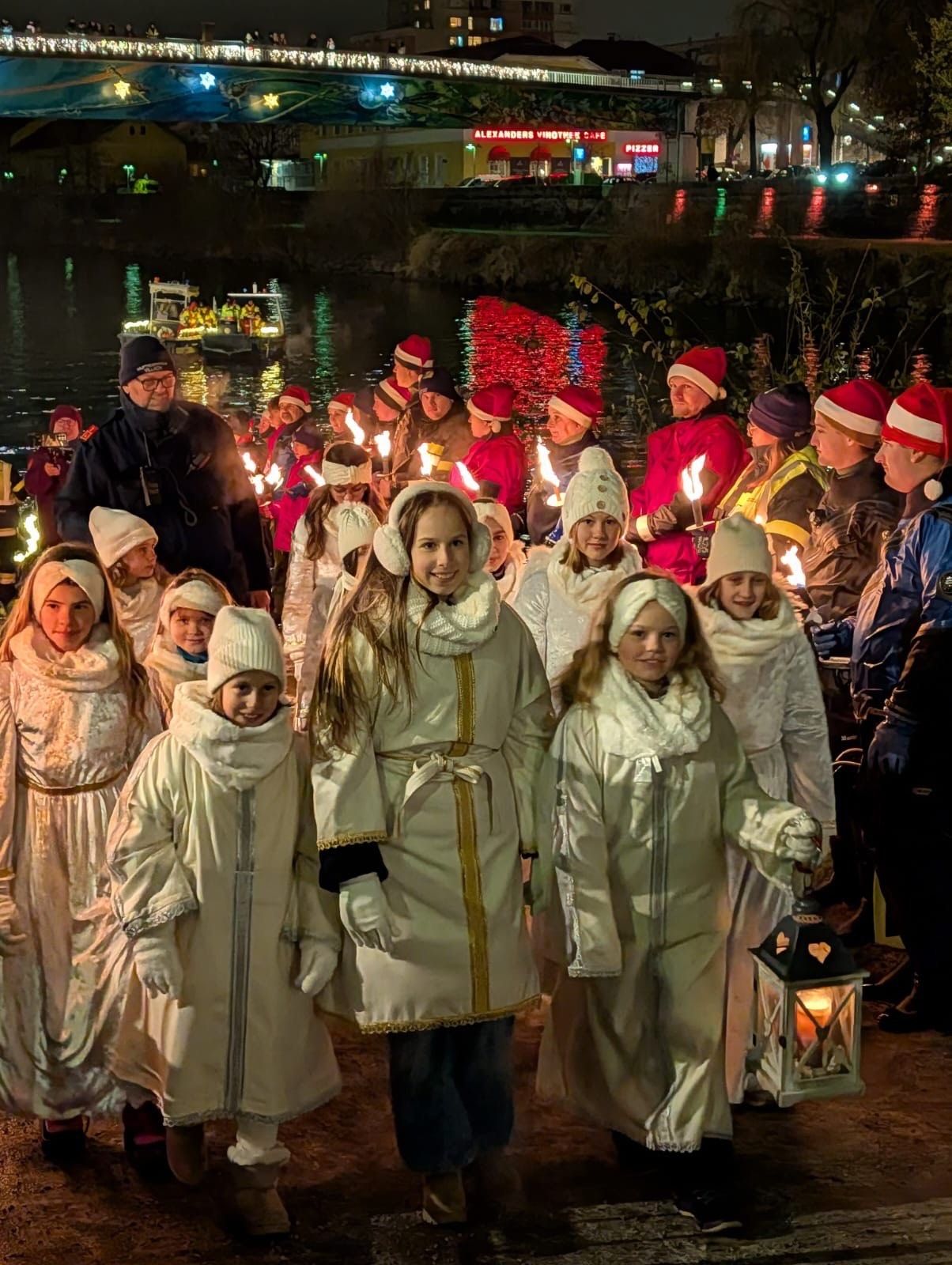 Eine Gruppe von Kindern in traditioneller Winterkleidung, die brennende Laternen halten, gehen bei Nacht auf einer verschneiten Straße. Im Hintergrund fährt ein Boot mit beleuchteten Lichtern auf dem Fluss.