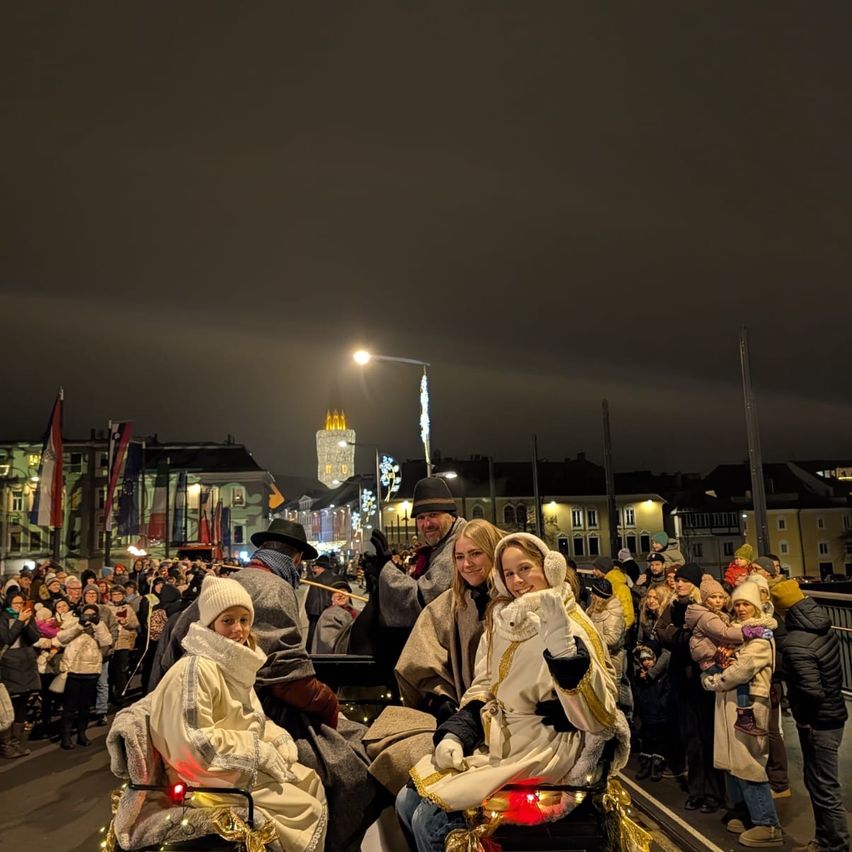 Eine Gruppe von Menschen in Winterkleidung sieht bei Nacht eine Parade. Zwei Erwachsene fahren in einer geschmückten Kutsche mit einem jungen Mädchen. Es gibt viele Zuschauer auf der Straße. Im Hintergrund sind Gebäude zu sehen.