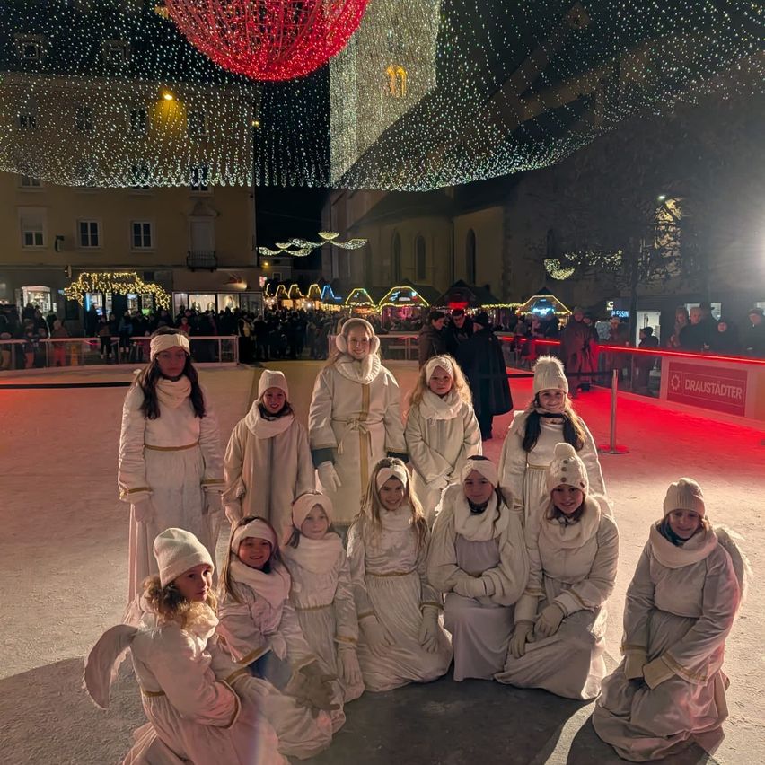Eine Gruppe junger Mädchen in weißen Kleidern posiert für ein Foto auf einer Eisbahn bei Nacht, geschmückt mit festlichen Lichtern und Ornamenten.