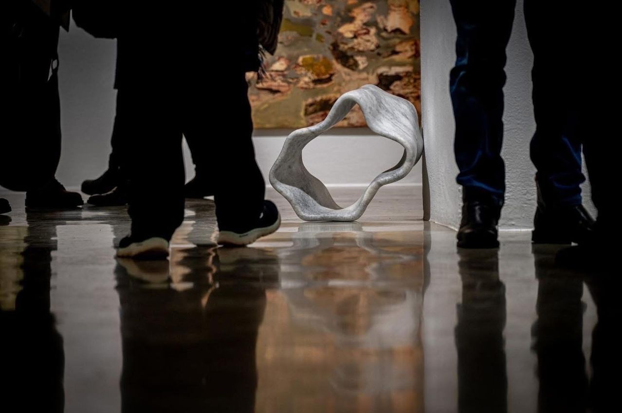 Zwei Personen gehen in einem Museum. Eine weiße Skulptur liegt auf dem Boden. Der Boden reflektiert ihre Schatten. Ein Gemälde ist an der Wand.