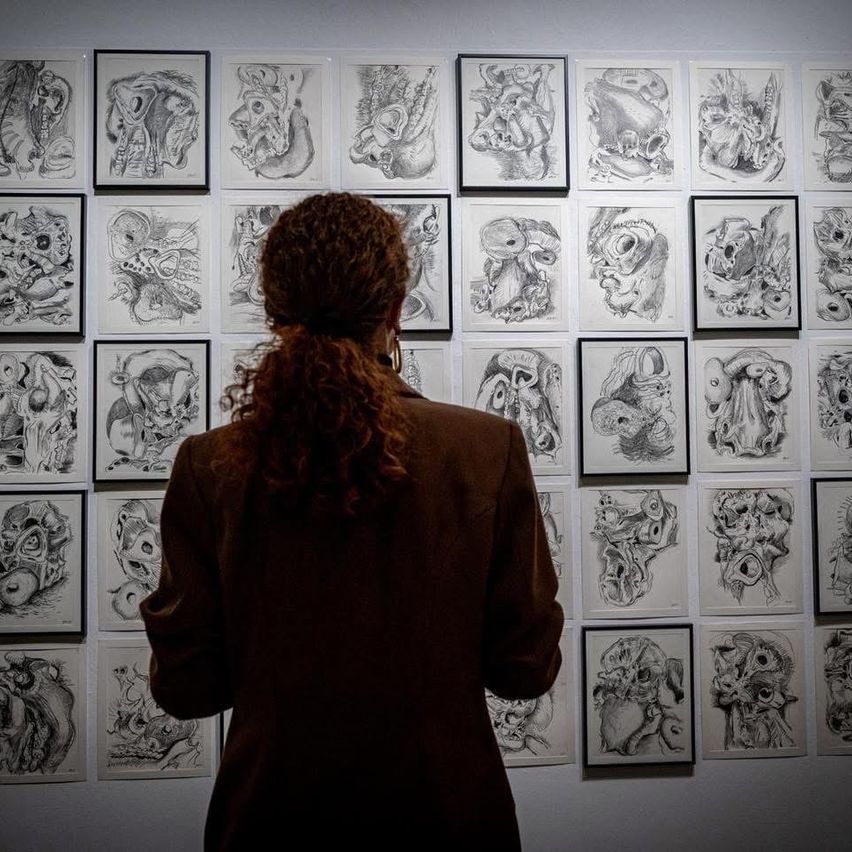 Eine Person mit lockigem Haar, die einen braunen Mantel trägt, steht vor einer Wand, die mit verschiedenen gerahmten Zeichnungen bedeckt ist. Die Zeichnungen zeigen menschliche und tierische Formen in Schwarz und Weiß.