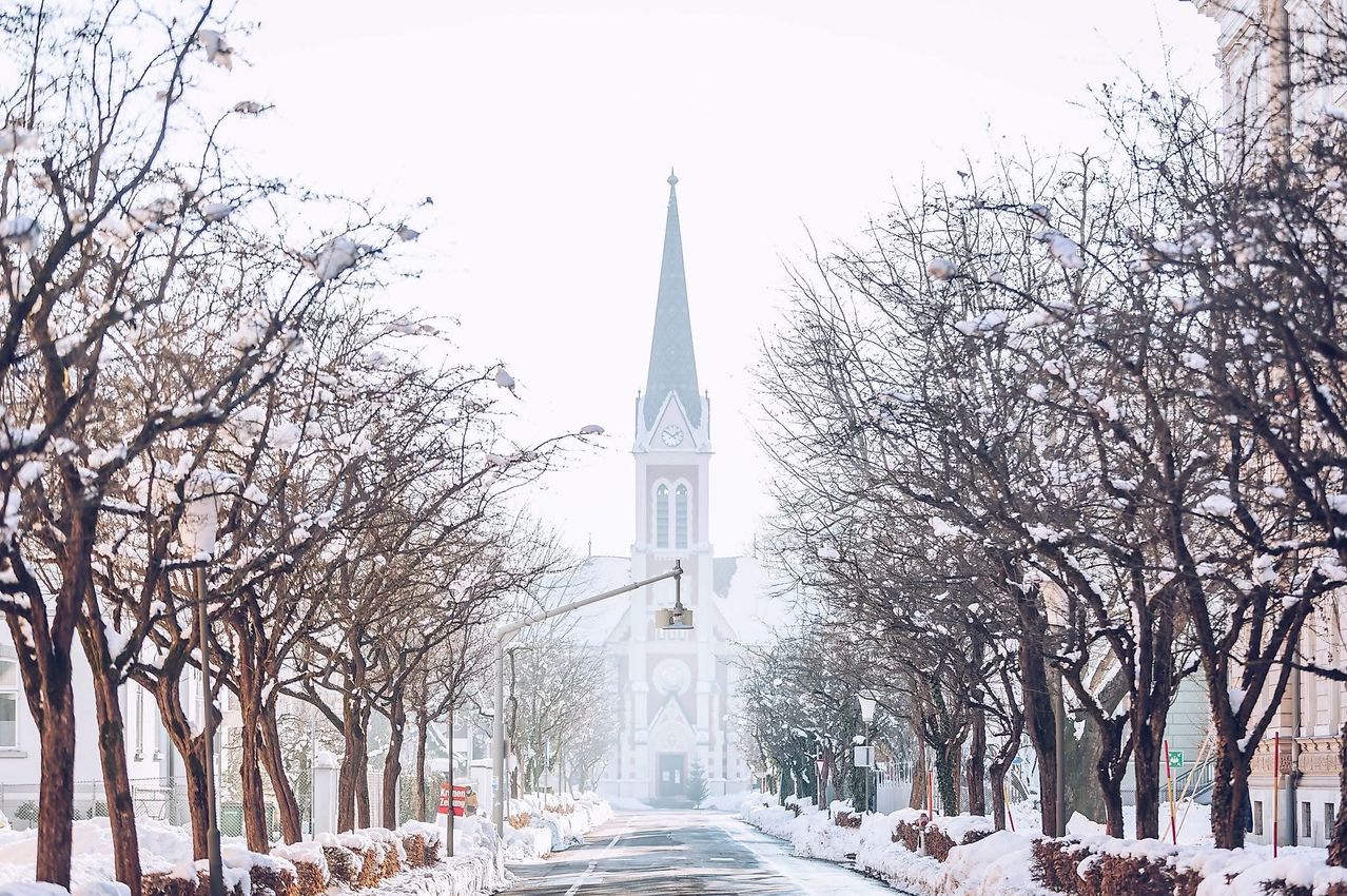 Eine verschneite Straße mit Bäumen und einer Kirche mit Turm und Uhrenturm in der Ferne. Schnee bedeckt den Boden und die Bäume.
