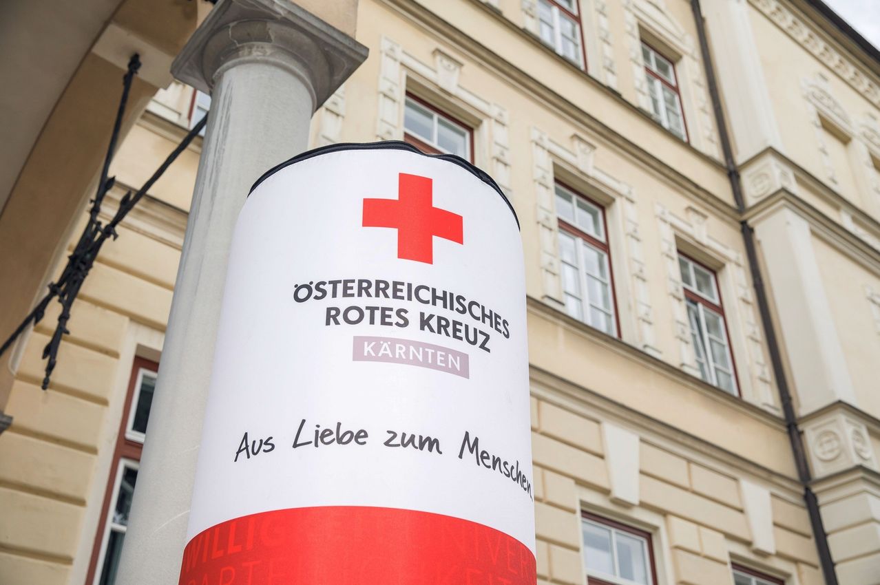 Ein rotes Kreuzbanner steht vor einem beigen Gebäude mit Fenstern. Es steht auf Deutsch geschrieben 'Aus Liebe zum Menschen'.