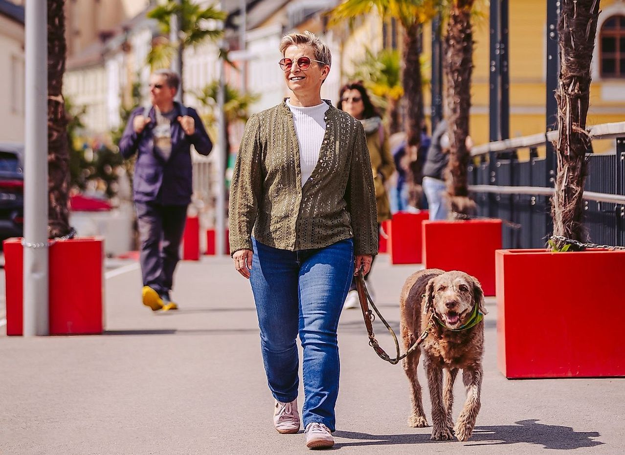 Eine ältere Frau mit Sonnenbrille führt ihren Hund auf einer Stadtstraße spazieren. Eine weitere Person läuft hinter ihr. Gebäude und Palmen säumen die Straße.