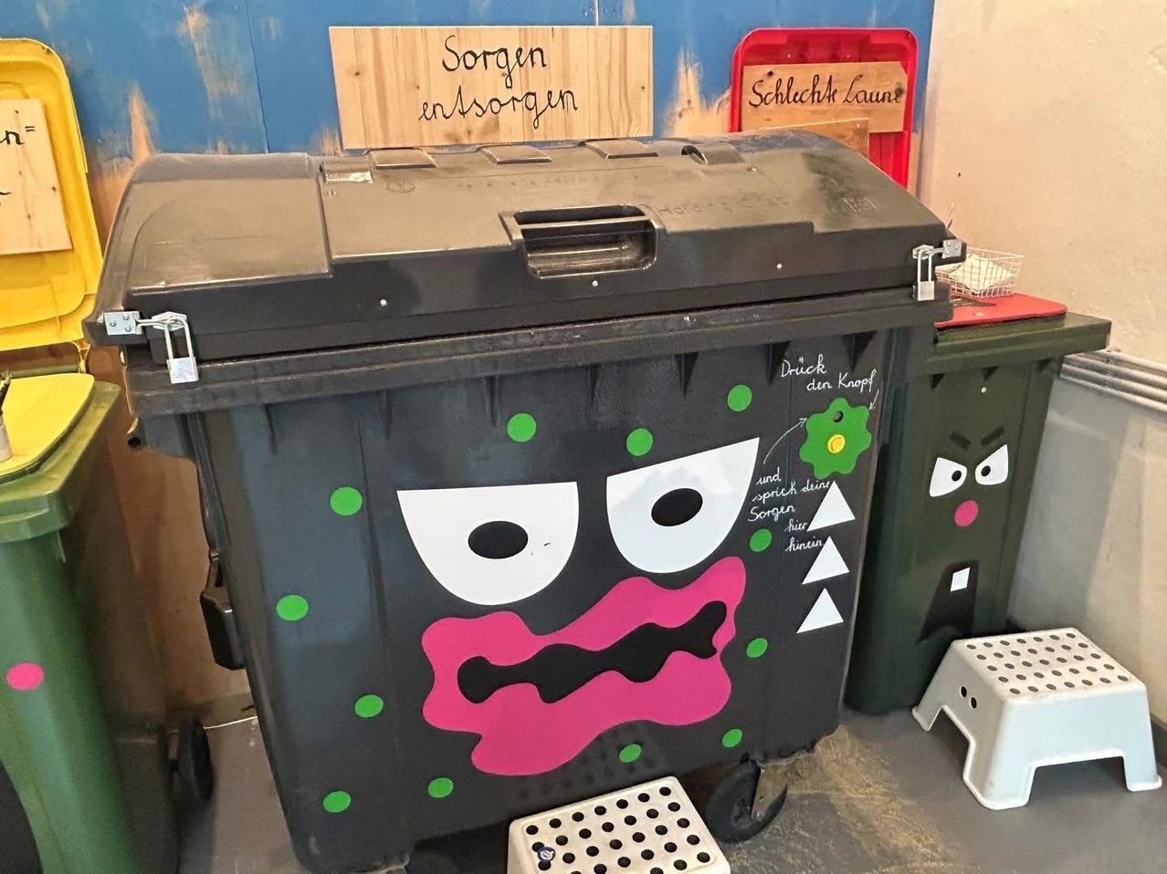 Ein schwarzer Mülleimer mit bemaltem Gesicht, beschriftet mit 'Knopf drücken'. Er hat grüne Punkte und einen rosa Mund. In der Nähe befindet sich ein Holzschild mit der Aufschrift 'Sorgen einsorgern'.
