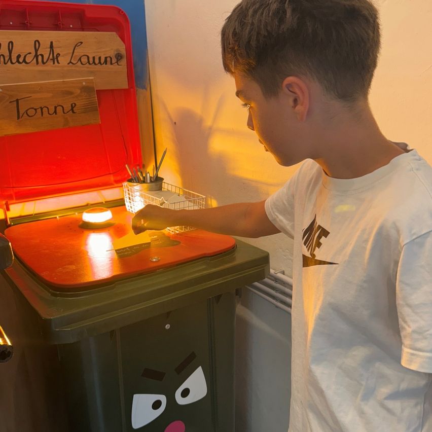 Ein Junge steht neben einem Mülleimer und schaut auf ein leuchtendes Licht darin. Er trägt ein weißes T-Shirt. Über dem Mülleimer steht ein Schild mit der Aufschrift 'SchlechkA laune Tonne'.