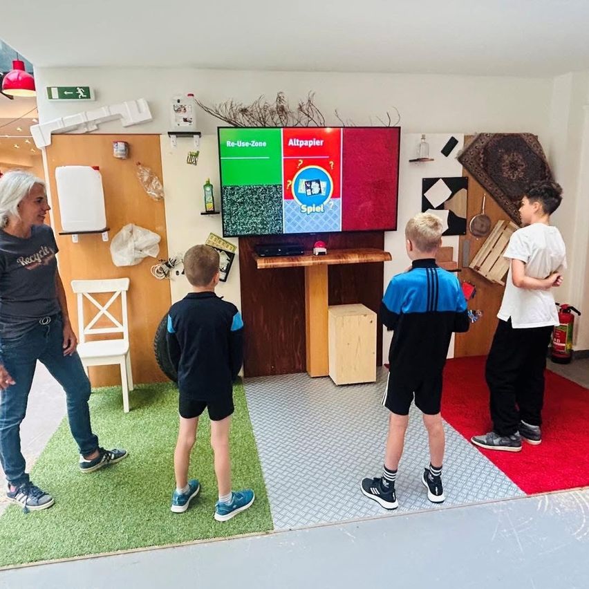Vier Jungen spielen ein Spiel in einem Raum mit einem großen Bildschirm. Ein Junge trägt ein blaues Shirt und Turnschuhe. Ein anderer Junge trägt ein schwarzes Shirt und Shorts. Eine Frau steht vor ihnen.