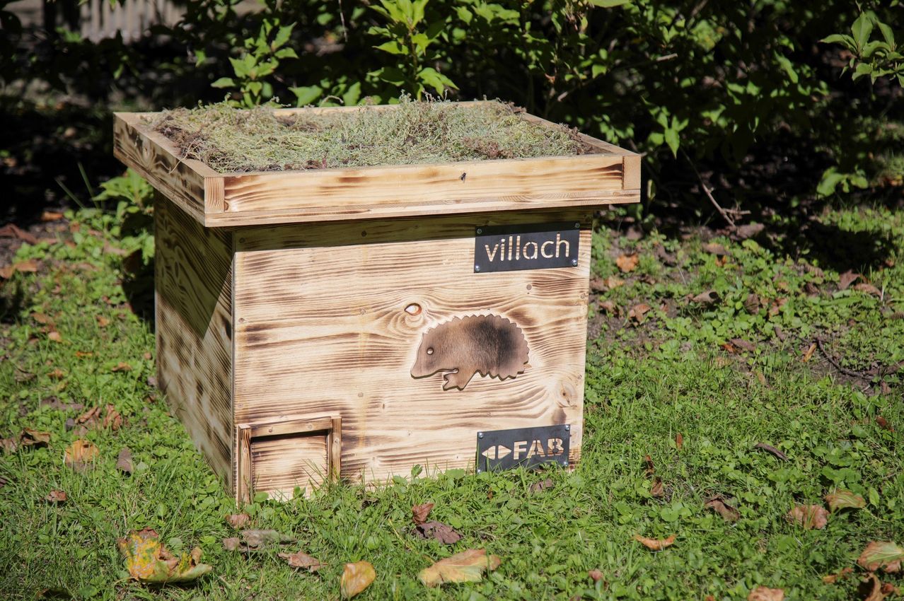 Eine Holzkiste mit einem Igeldesign und einem Schild mit den Aufschriften 'villach' und 'FAB' steht auf Gras mit verstreuten Blättern.