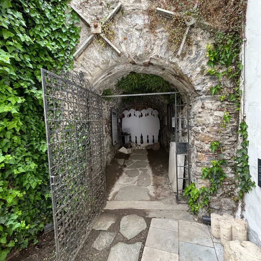 Ein Bogengang mit Efeu bewachsenen Steinmauern führt zu einem Tunnel mit einem eisernen Tor und einem Steinweg. Im Inneren befindet sich ein Raum mit einer Ausstellung weißer Figuren und Weinflaschen.