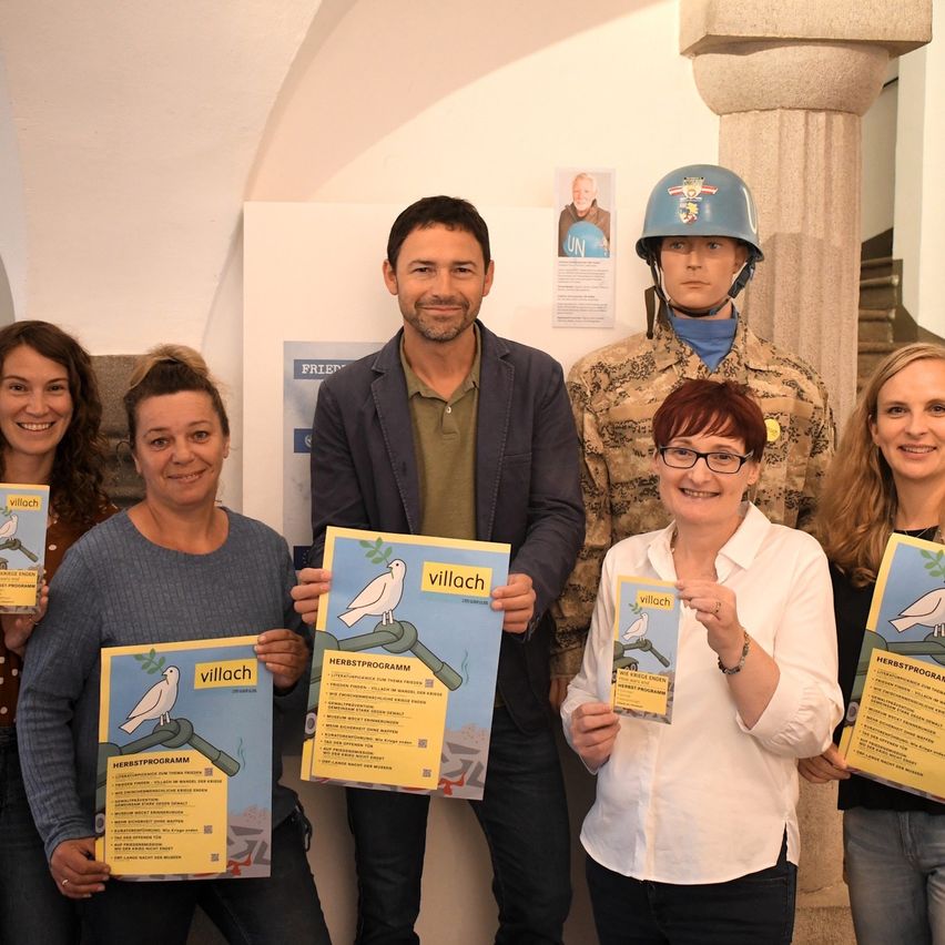 Eine Gruppe von Erwachsenen hält Flyer mit einem Vogel und dem Wort 'Villach' darauf. Sie stehen vor einer Wand mit einem Plakat und einer Puppe mit Helm. Dahinter steht eine Säule.