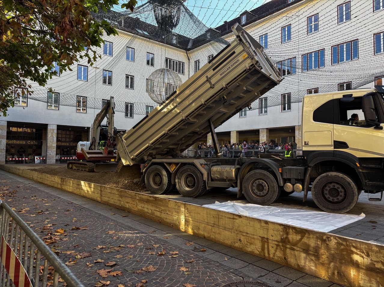 Ein Lastwagen entlädt seine Fracht vor einem großen Gebäude, neben einem Kran und mit vielen zuschauenden Menschen.
