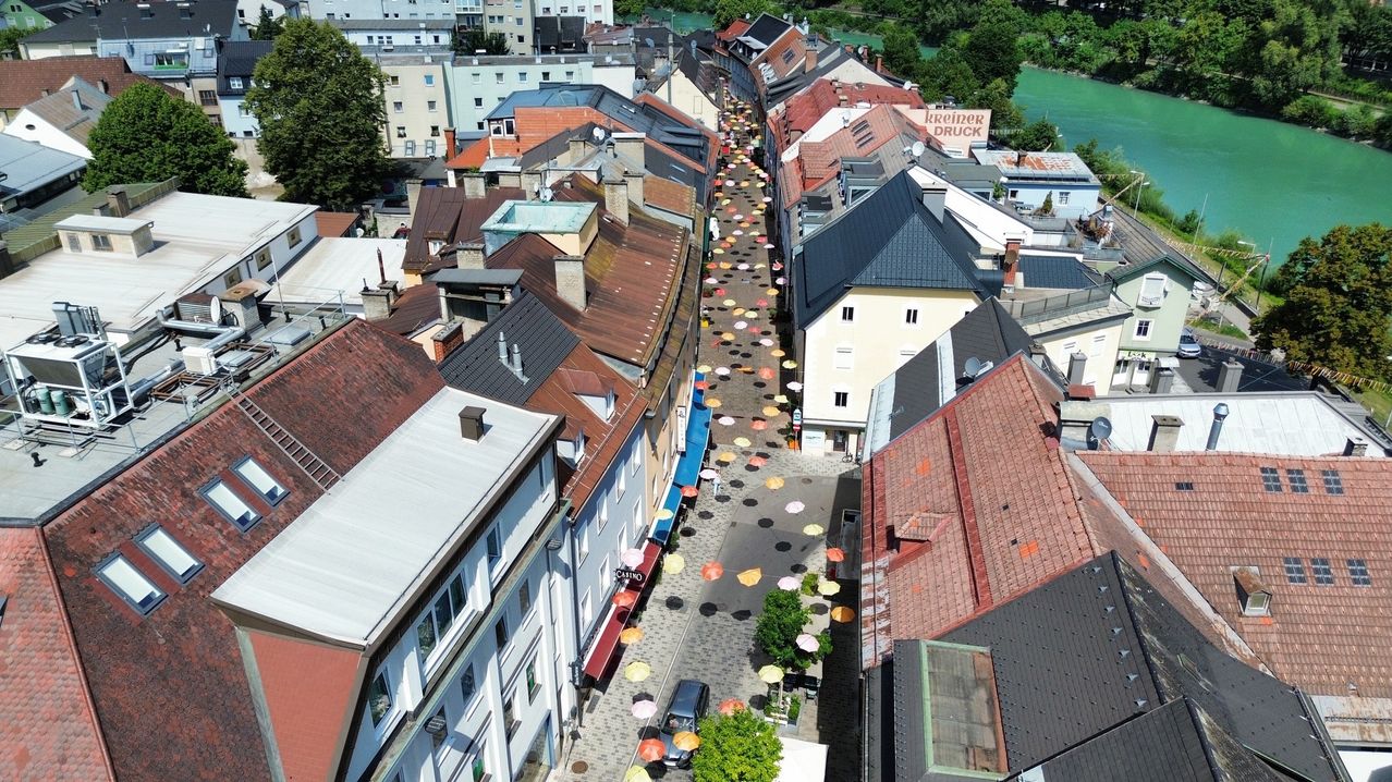 Luftaufnahme einer Straße mit vielen bunten Regenschirmen auf dem Boden, gesäumt von Gebäuden mit unterschiedlich gefärbten Dächern. In der Ferne befindet sich ein Fluss und Bäume umgeben das Gebiet.
