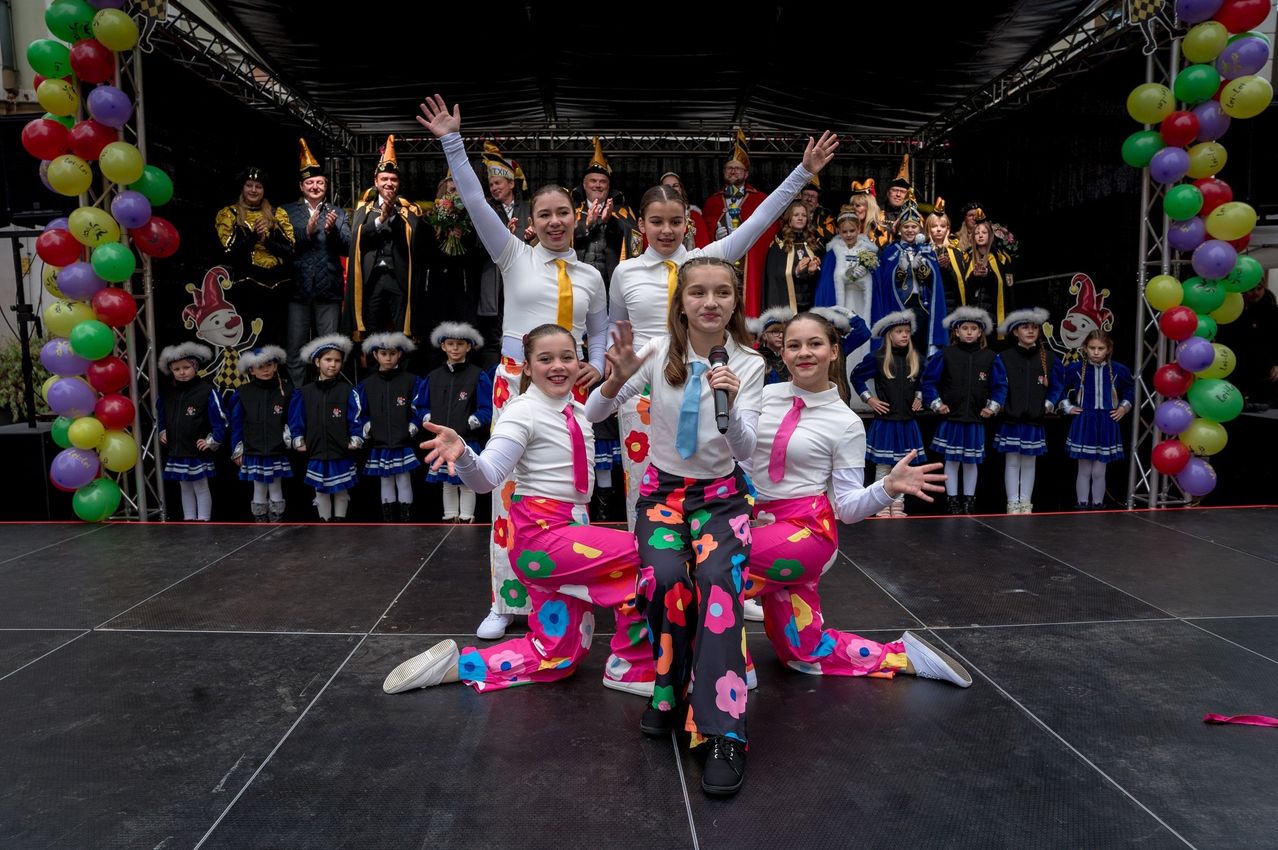 Vier Mädchen in weißen Oberteilen und bunten Hosen treten mit Mikrofonen auf der Bühne auf. Dahinter applaudiert eine Menschenmenge in Kostümen.