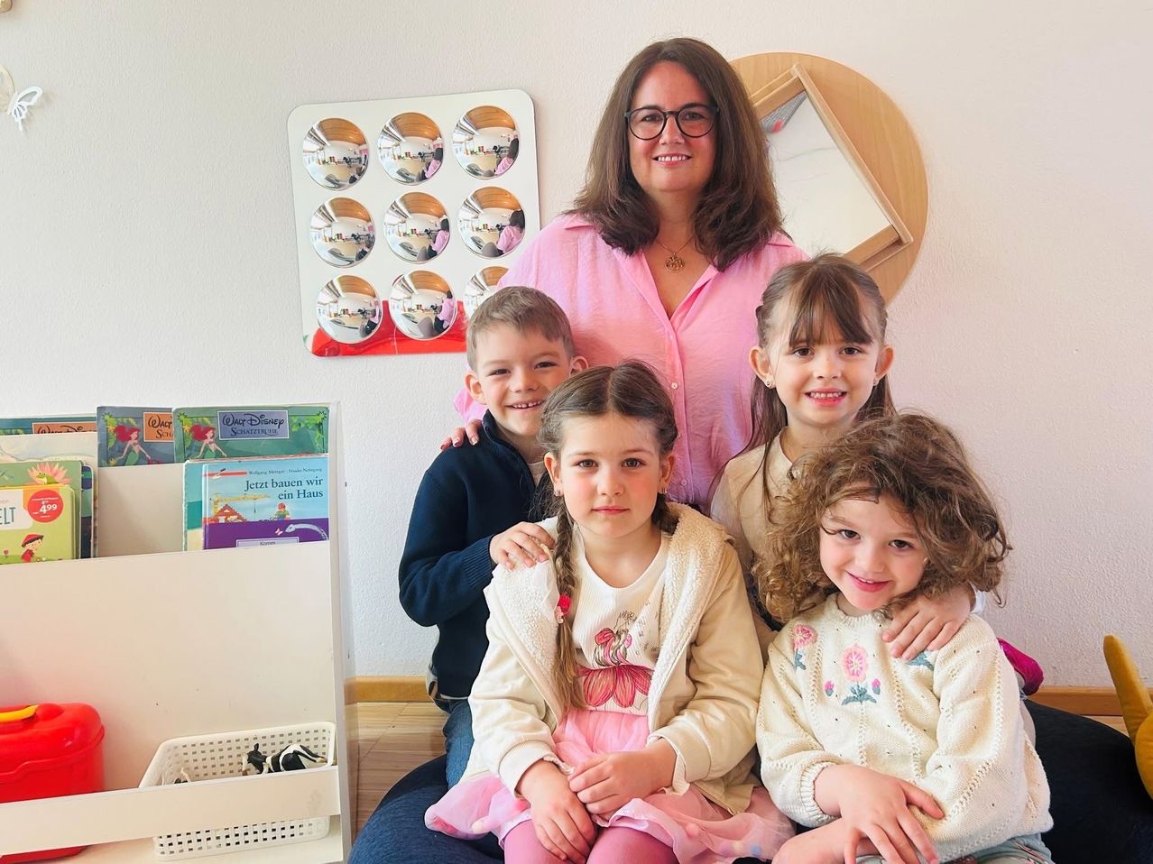 Eine Frau in einem rosa Hemd ist mit vier Kindern, drei Mädchen und einem Jungen. Die Kinder lächeln und schauen in die Kamera. Die Frau steht hinter ihnen. Der Raum hat eine Wand mit einem Spiegel und einem Regal mit Büchern.