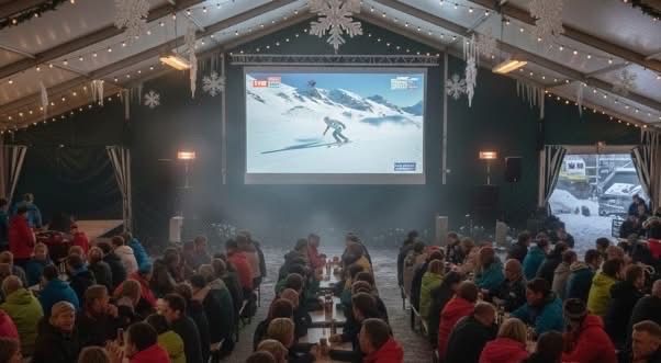 Eine Menschenmenge sitzt in einem Zelt um Tische herum und schaut sich einen Skiwettbewerb auf einer großen Leinwand an. Schneeflocken hängen von der Decke und es herrscht eine festliche Atmosphäre.